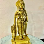 Gold-toned Hindu deity statue in ornate armor and crown, holding a staff, placed on a marble surface with 'HANDICRAFTS' visible at the base and a brand logo in the corner.  thebrasshandicrafts.com