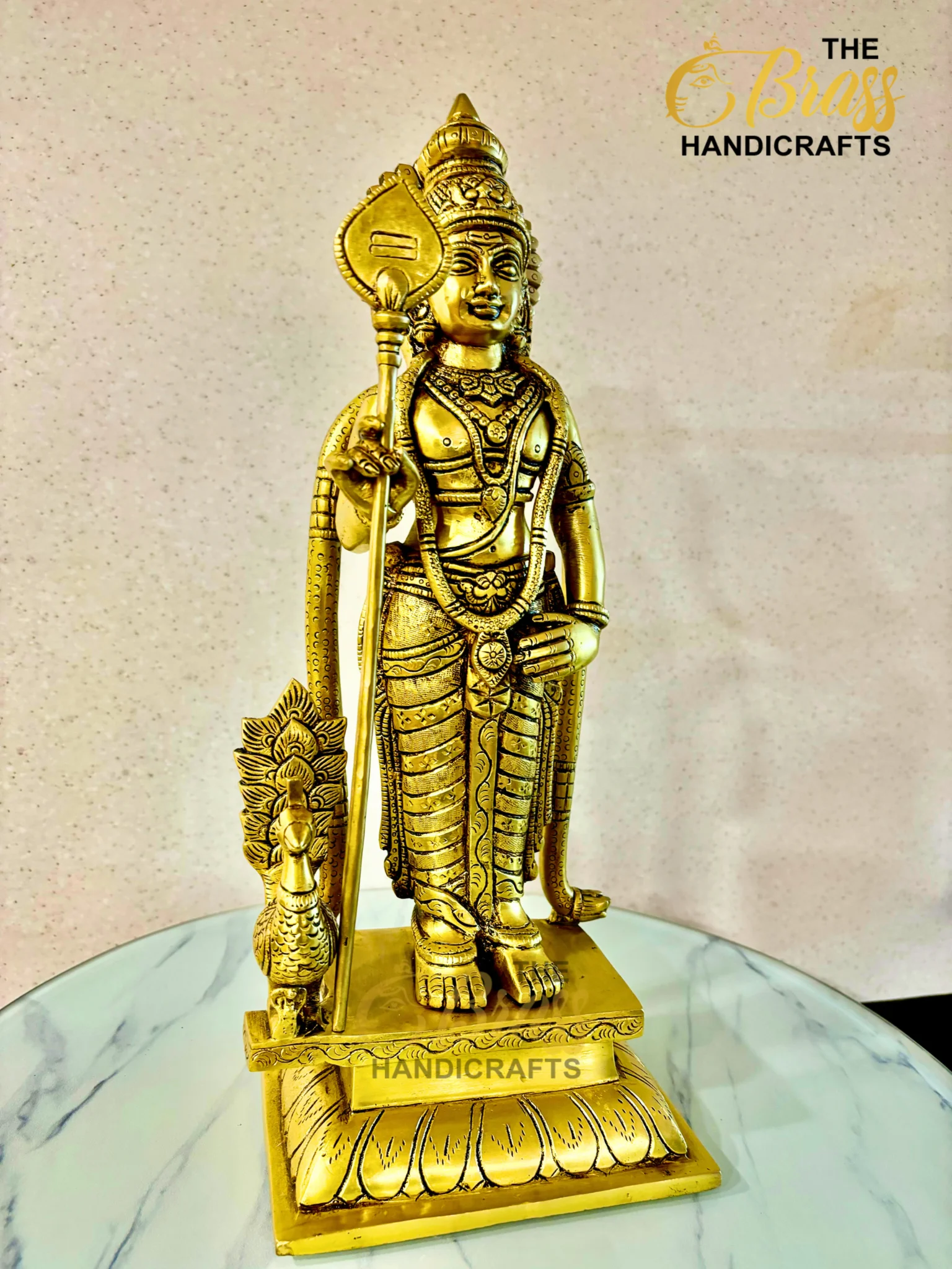 Gold-toned Hindu deity statue in ornate armor and crown, holding a staff, placed on a marble surface with 'HANDICRAFTS' visible at the base and a brand logo in the corner.  thebrasshandicrafts.com