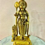 Golden brass statue of a Hindu deity standing with a staff, displayed on a marble-topped surface with a decorative base and a 'HANDICRAFTS' plaque visible at the bottom.  thebrasshandicrafts.com