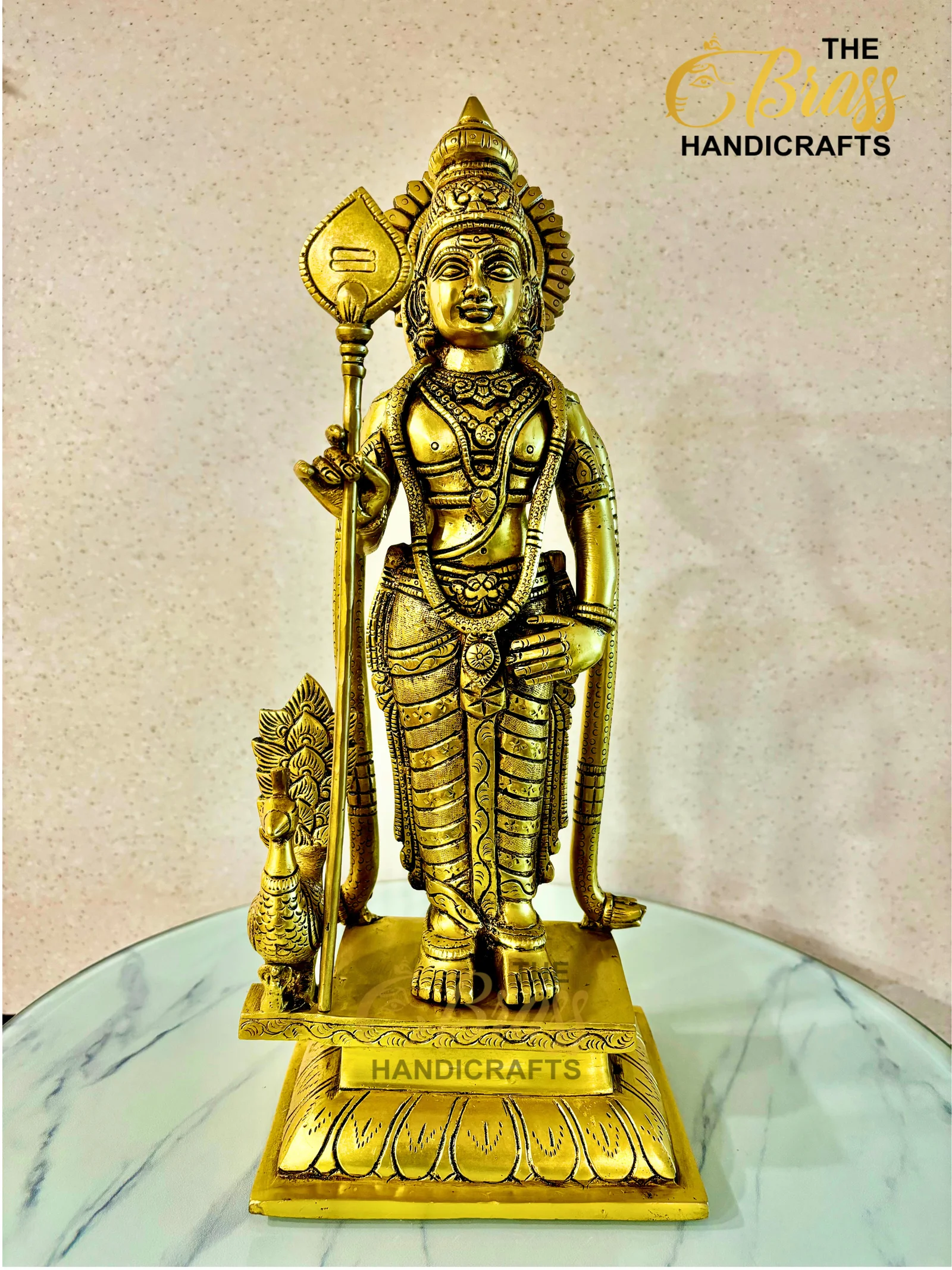 Golden brass statue of a Hindu deity standing with a staff, displayed on a marble-topped surface with a decorative base and a 'HANDICRAFTS' plaque visible at the bottom.  thebrasshandicrafts.com