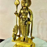 Gold-colored statue of a Hindu deity holding a staff, richly ornamented and standing on a marble base with a decorative frame.  thebrasshandicrafts.com