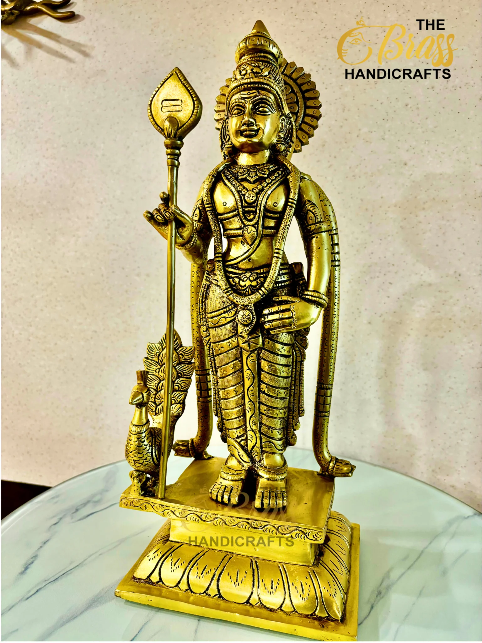 Gold-colored statue of a Hindu deity holding a staff, richly ornamented and standing on a marble base with a decorative frame.  thebrasshandicrafts.com