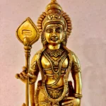 Gold-toned Hindu deity statue with ornate jewelry and a tall staff, close-up view on a light speckled background.  thebrasshandicrafts.com