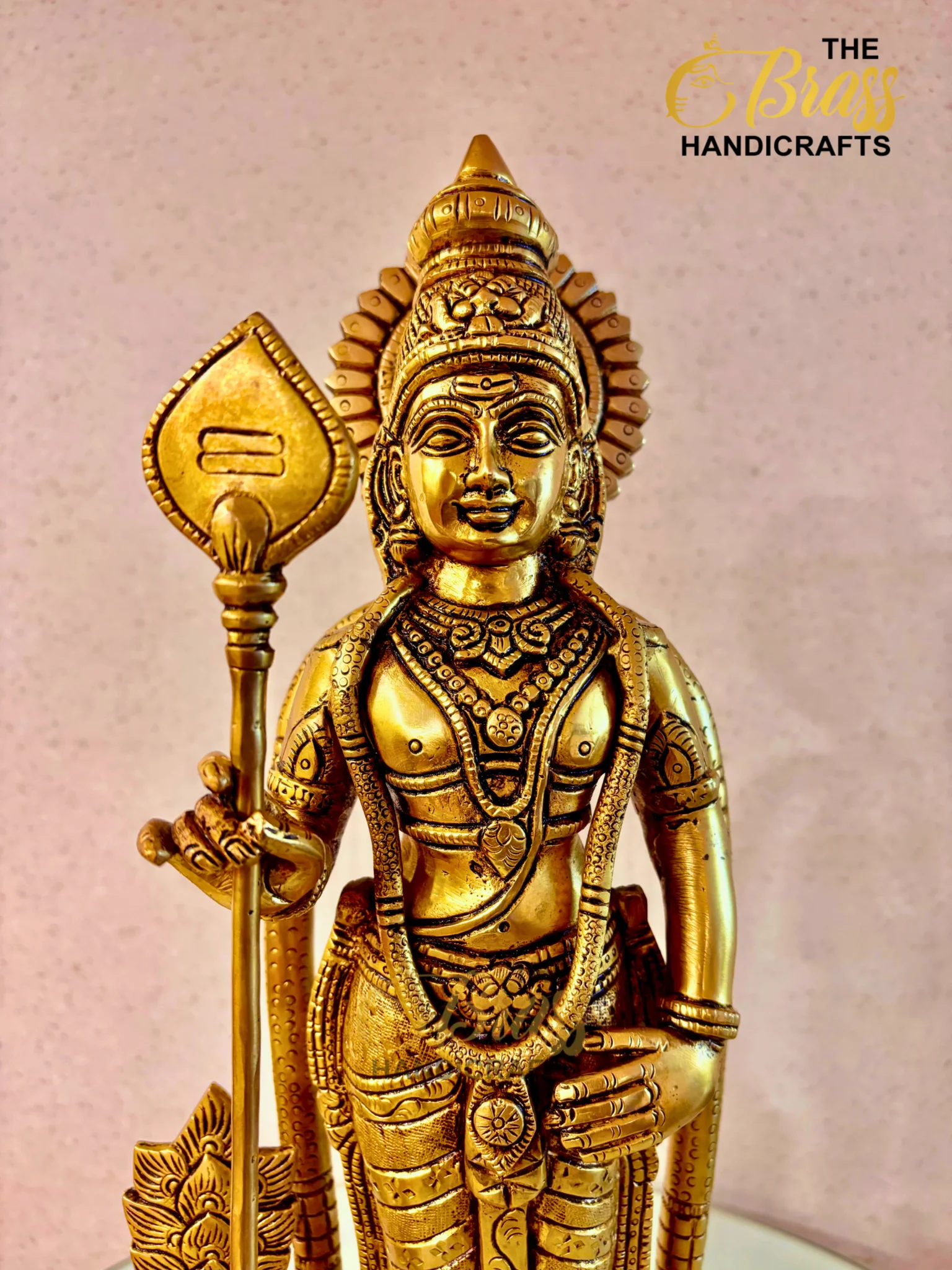 Gold-toned Hindu deity statue with ornate jewelry and a tall staff, close-up view on a light speckled background.  thebrasshandicrafts.com