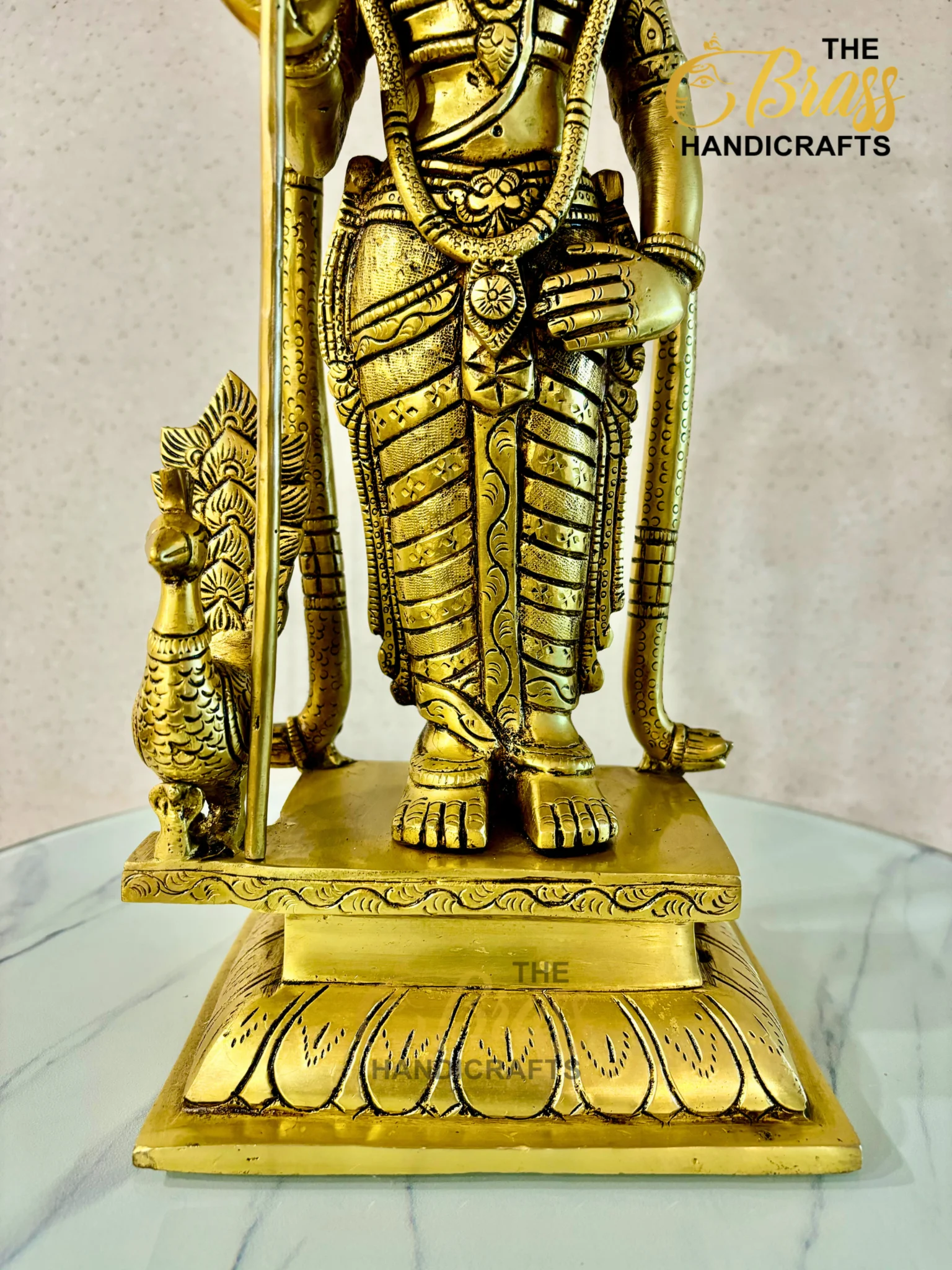 Close-up of a gold brass statue's lower torso, hands, and ornate belt, standing on a carved pedestal with decorative patterns; watermark 'The Brass Handicrafts' visible in the corner.  thebrasshandicrafts.com