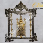 Ornate metal frame housing a golden Hindu deity statue adorned with jewelry and hanging ornaments, set against a gray backdrop.  thebrasshandicrafts.com