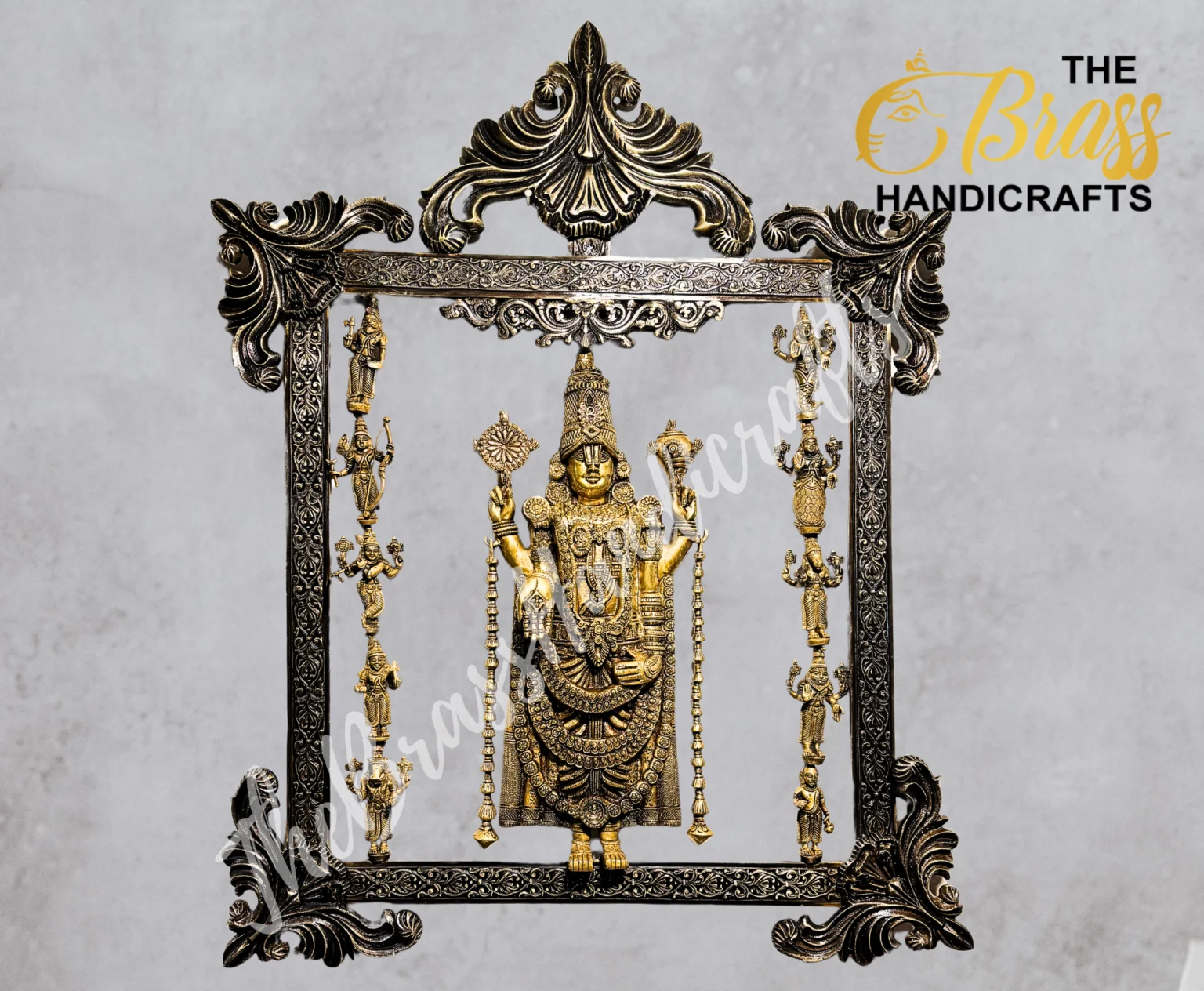 Ornate metal frame housing a golden Hindu deity statue adorned with jewelry and hanging ornaments, set against a gray backdrop.  thebrasshandicrafts.com