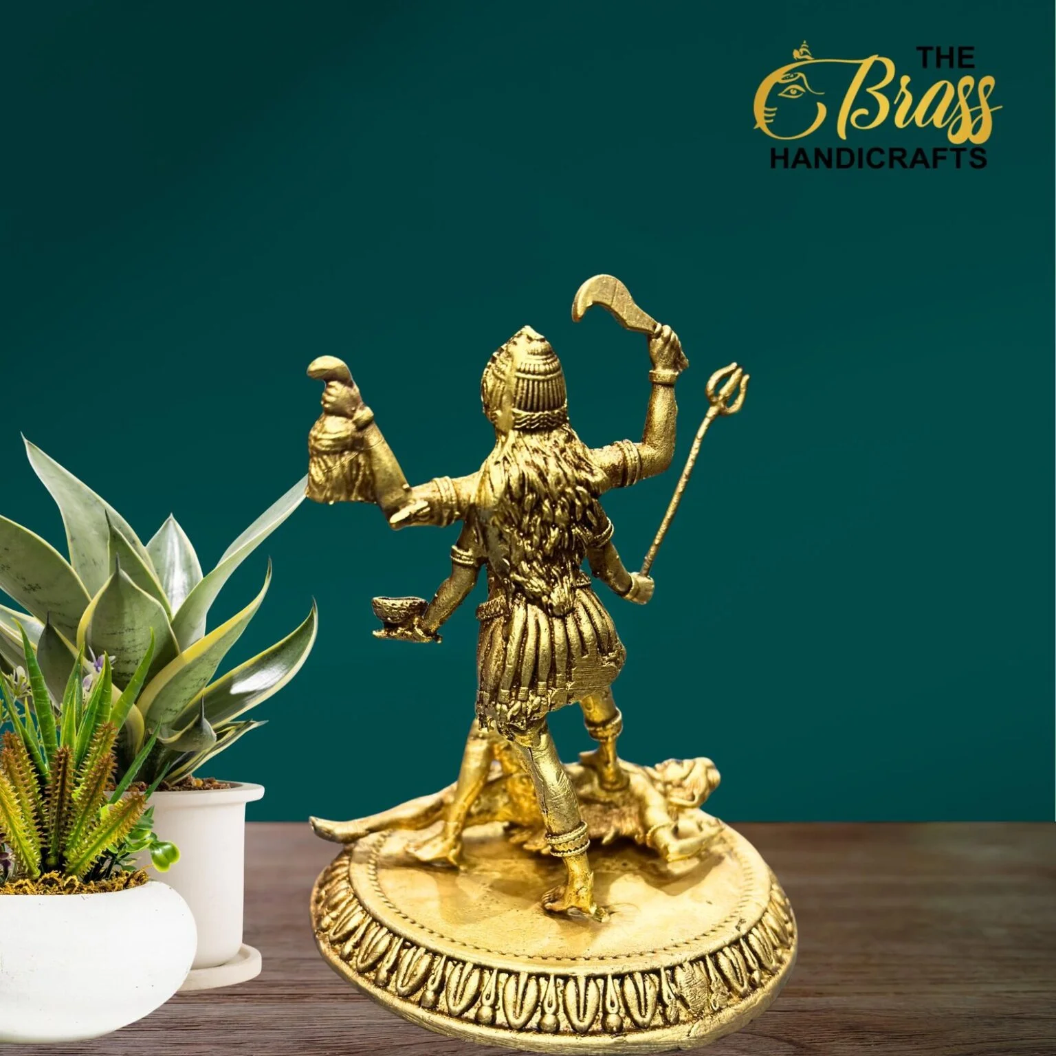 Gold brass statue of Shiva as Nataraja with raised arms, on a decorative base beside a potted plant against a teal background.  thebrasshandicrafts.com