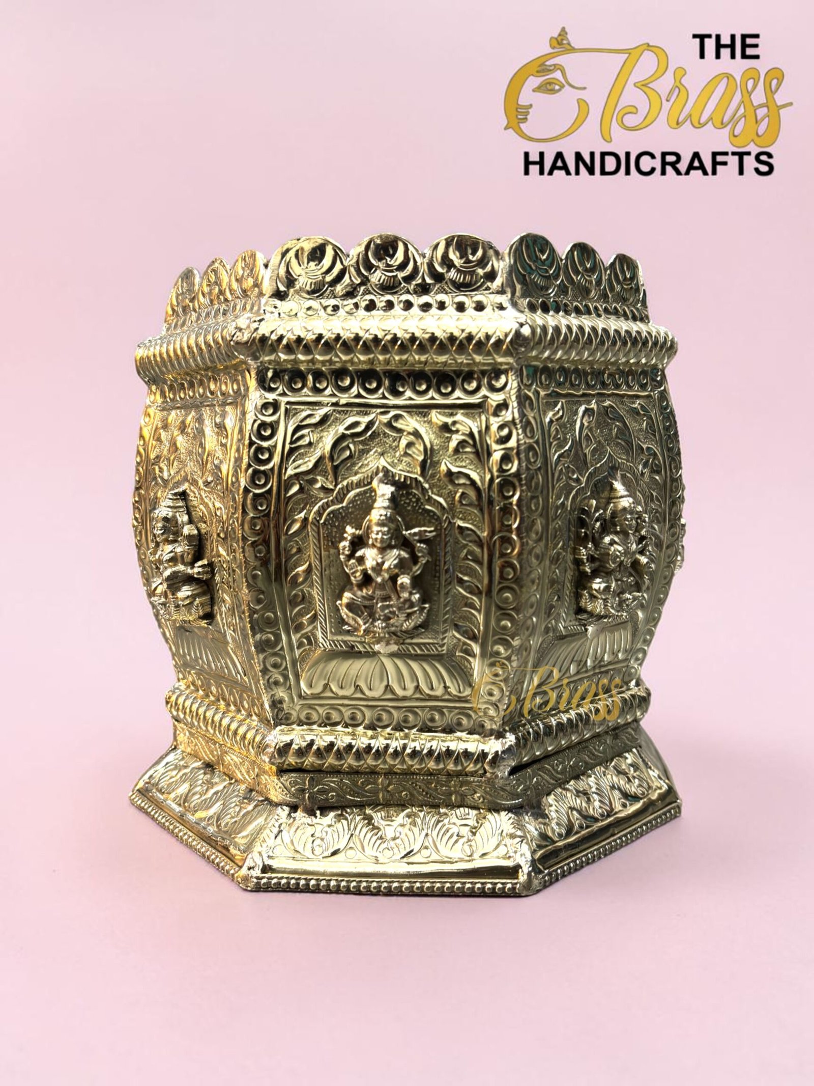 Gold ornate brass shrine with Buddha reliefs and intricate patterns on a pink background.  thebrasshandicrafts.com