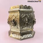 Ornate silver metal decorative shrine with embossed panels and small figurines, set against a pink background.  thebrasshandicrafts.com
