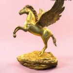 Gold Pegasus statue with wings raised, mounted on a rocky gold pedestal against a pink background, branding from The Brass Handicrafts in the corner.  thebrasshandicrafts.com