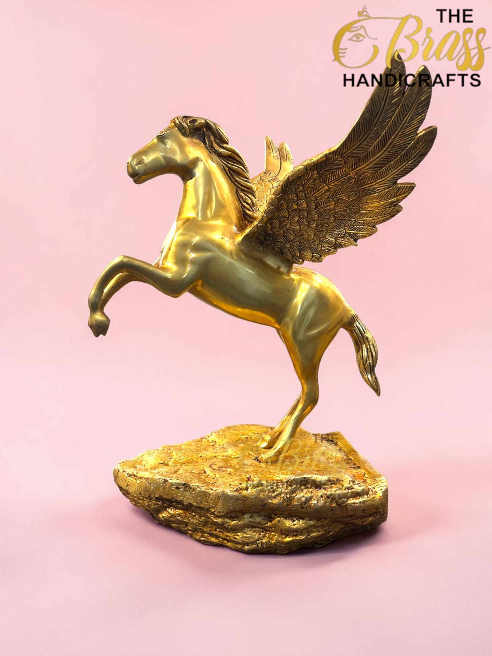 Gold Pegasus statue with wings raised, mounted on a rocky gold pedestal against a pink background, branding from The Brass Handicrafts in the corner.  thebrasshandicrafts.com