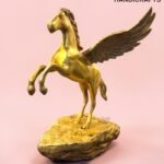 Golden Pegasus statue with wings raised, standing on a rocky base against a pink background.  thebrasshandicrafts.com