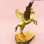 Gold-painted Pegasus statue with outstretched wings perched on a textured gold pedestal, pink background with a branding watermark in the corner.  thebrasshandicrafts.com
