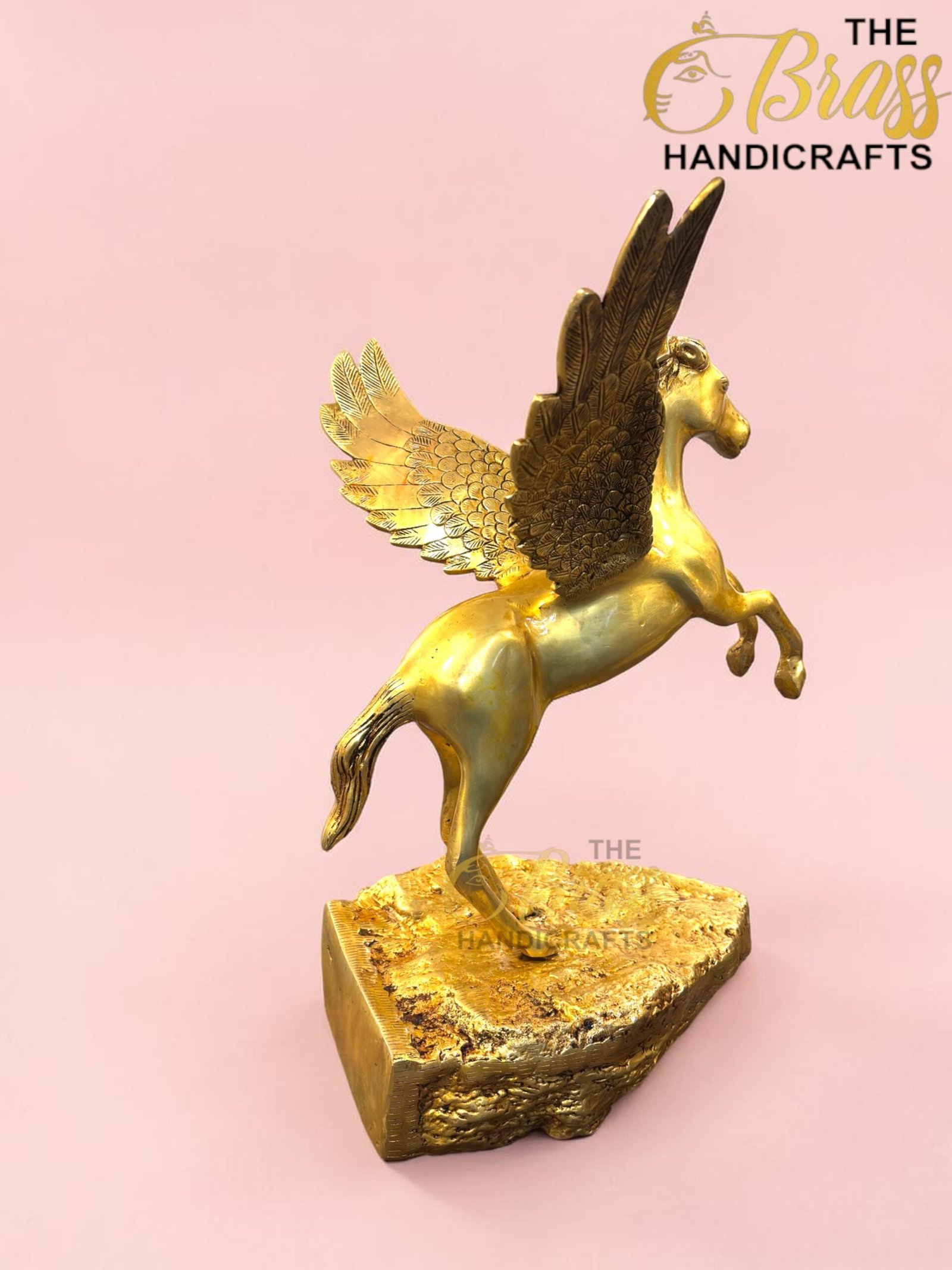 Gold-painted Pegasus statue with outstretched wings perched on a textured gold pedestal, pink background with a branding watermark in the corner.  thebrasshandicrafts.com
