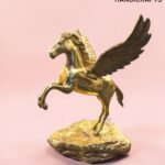Gold Pegasus statue with wings on a rocky base against a pink background, featuring The Brass Handicrafts logo in the corner.  thebrasshandicrafts.com