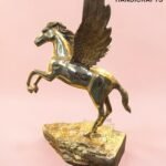 Gold-toned Pegasus statue with spread wings perched on a textured rock base against a pink background, a brass handicraft.  thebrasshandicrafts.com