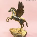 Brass gold-toned statue of a winged horse (Pegasus) rearing on a rocky base against a pink backdrop.  thebrasshandicrafts.com