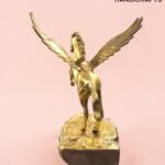 Gold statue of a winged horse (Pegasus) rearing on a textured pedestal against a pink background with a subtle logo watermark nearby.  thebrasshandicrafts.com