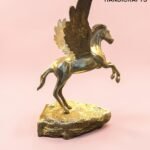 Golden Pegasus sculpture with spread wings perched on a rocky base against a pink background.  thebrasshandicrafts.com