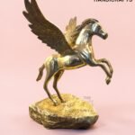 Gold Pegasus sculpture with wings spread, standing on a textured pedestal against a pink background; features 'The Brass Handicrafts' logo in the corner.  thebrasshandicrafts.com
