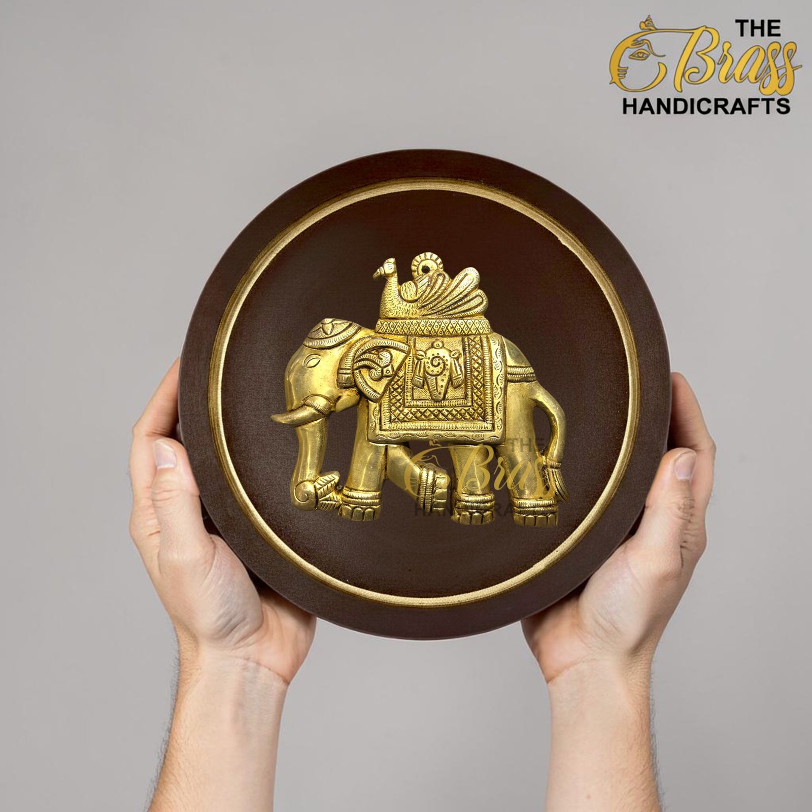 Hand holds a round brown plaque with a gold embossed elephant, logo partially visible in top right reading 'The Boss Handicrafts'  thebrasshandicrafts.com