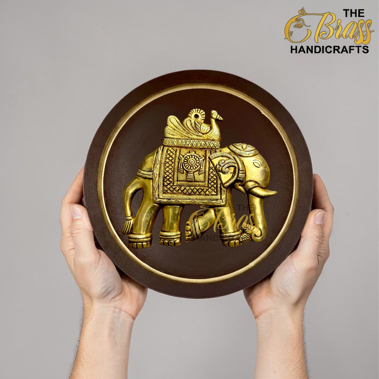 Person holding a round wooden shield with a gold embossed elephant design; brand logo in the top-right corner reads 'The Boss Handicrafts'.  thebrasshandicrafts.com