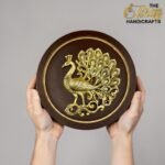 Two hands hold a round dark plaque featuring an ornate gold peacock design at the center; 'The Brass Handicrafts' logo appears in the top-right corner.  thebrasshandicrafts.com