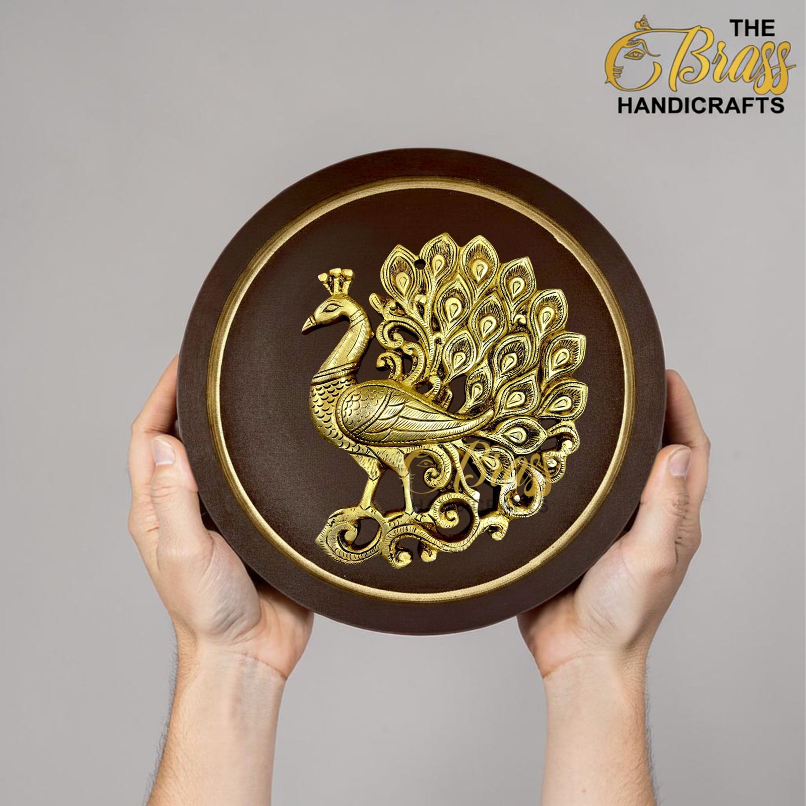 Two hands hold a round dark plaque featuring an ornate gold peacock design at the center; 'The Brass Handicrafts' logo appears in the top-right corner.  thebrasshandicrafts.com