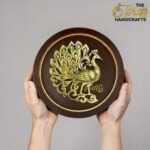 Brown circular plaque with an ornate gold peacock design being held by two hands in front of a gray background (watermark visible).  thebrasshandicrafts.com