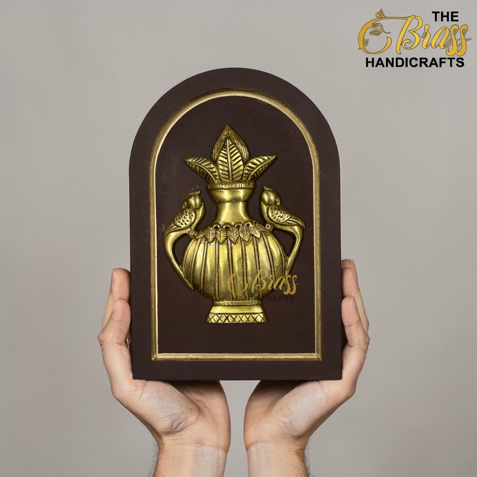 Person holding a dark brown, ornate plaque with a gold embossed emblem featuring a crown and leaf design; 'The Brass Handicrafts' logo is visible in the top right corner.  thebrasshandicrafts.com