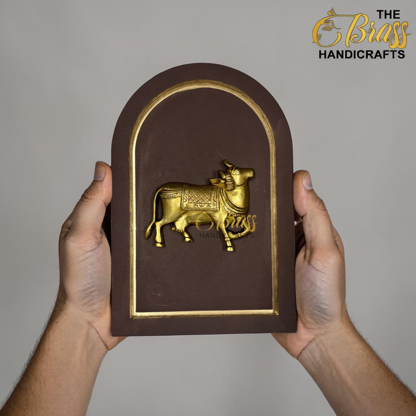 Hands hold a brown arch-shaped plaque with a gold embossed horse relief and a gold border against a gray background.  thebrasshandicrafts.com