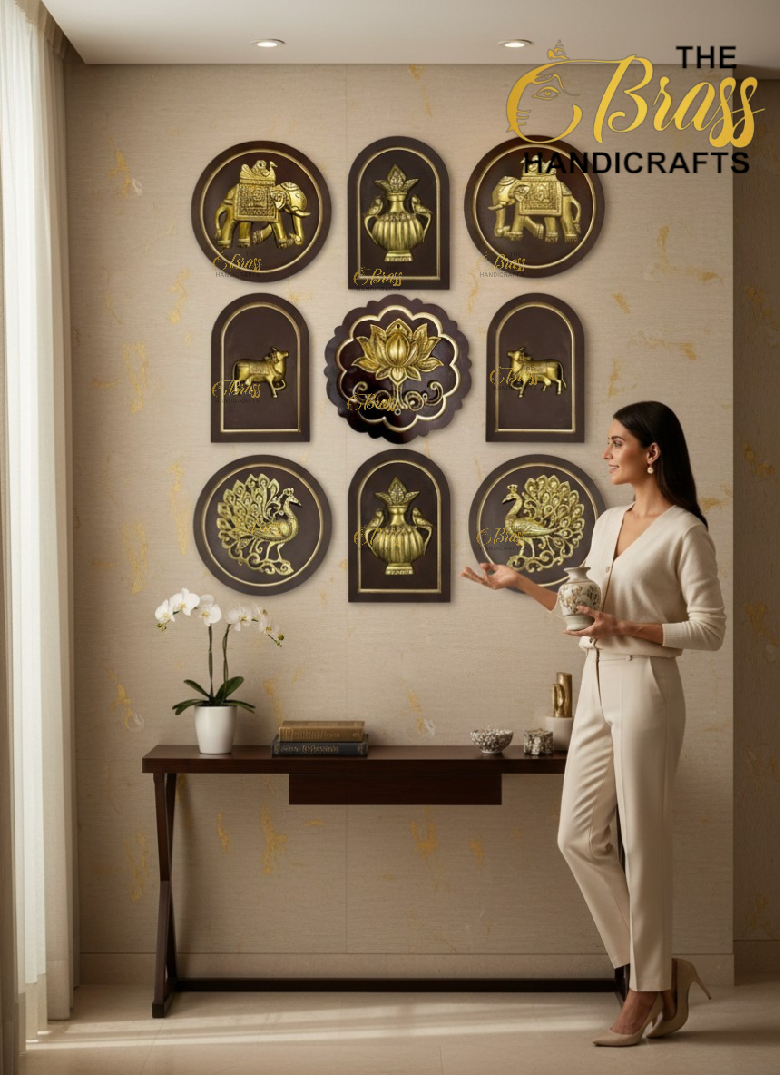 Showroom scene with gold embossed wall plaques (elephants, peacocks and lotus) on a beige wall, and a woman in a cream suit holding a vase next to the display, The Biass Handicrafts logo visible in the corner.  thebrasshandicrafts.com