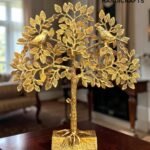 Gold-toned metal tree sculpture with two birds perched on leafy branches, displayed on a wooden table indoors (logo visible).  thebrasshandicrafts.com