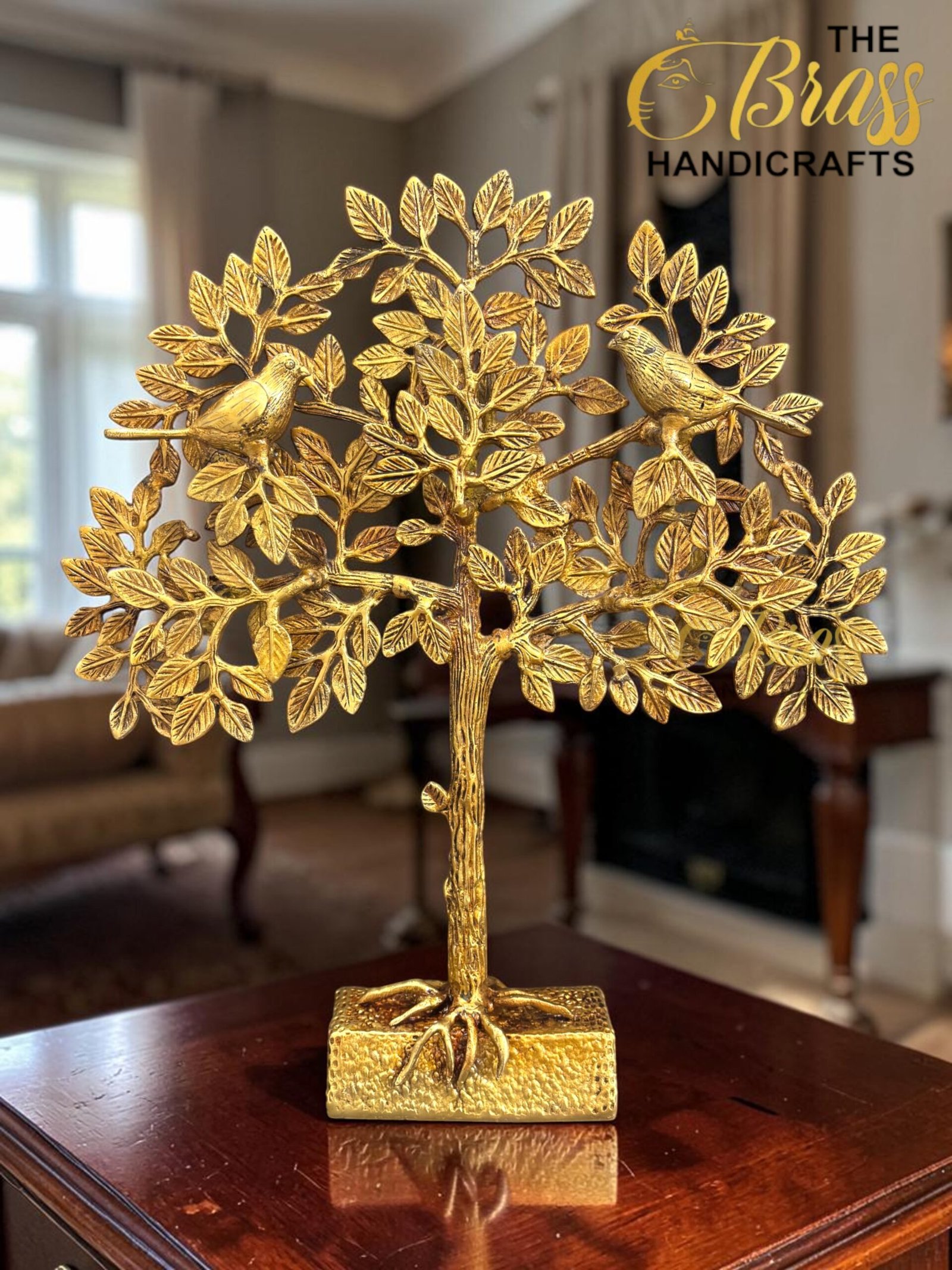 Gold-toned metal tree sculpture with two birds perched on leafy branches, displayed on a wooden table indoors (logo visible).  thebrasshandicrafts.com