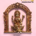 Gold-toned ornate statue of a Hindu goddess set in an intricate arch frame on a pink background with a 'The Brass Handicrafts' logo in the corner.  thebrasshandicrafts.com