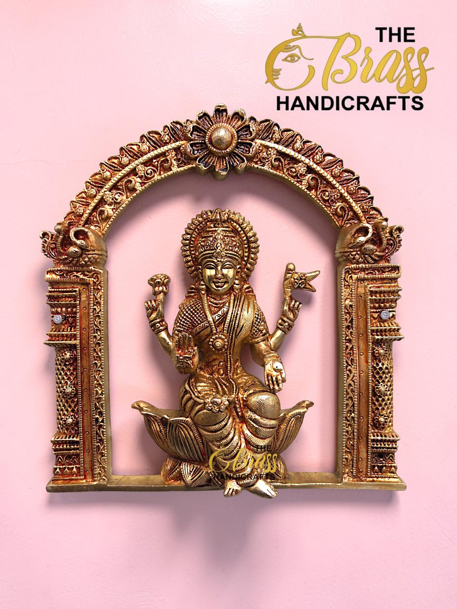 Gold-toned ornate statue of a Hindu goddess set in an intricate arch frame on a pink background with a 'The Brass Handicrafts' logo in the corner.  thebrasshandicrafts.com