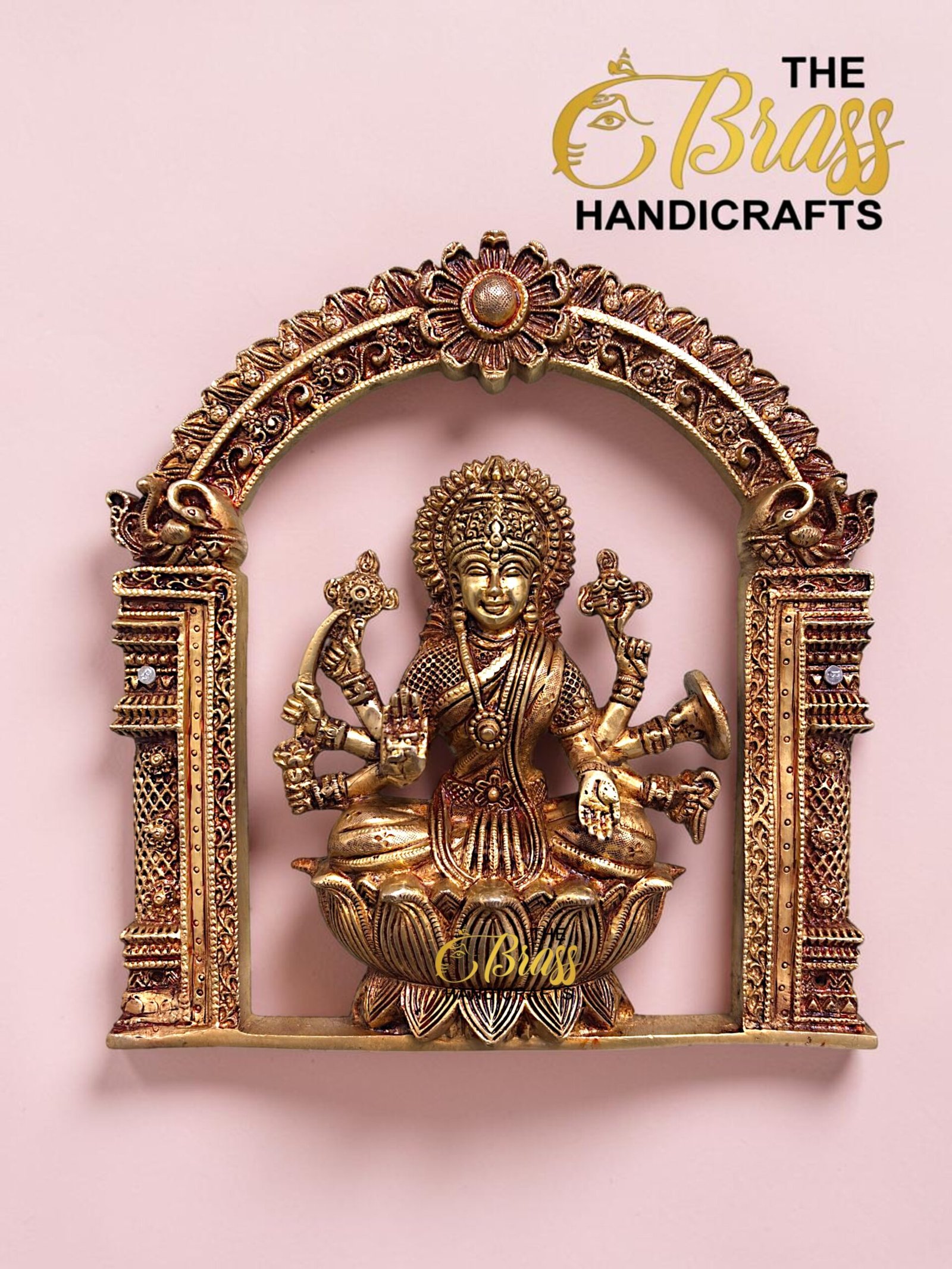 Ornate gold brass statue of a Hindu deity inside an intricate arch frame on a pink background, with 'The Brass Handicrafts' logo in the corner.  thebrasshandicrafts.com