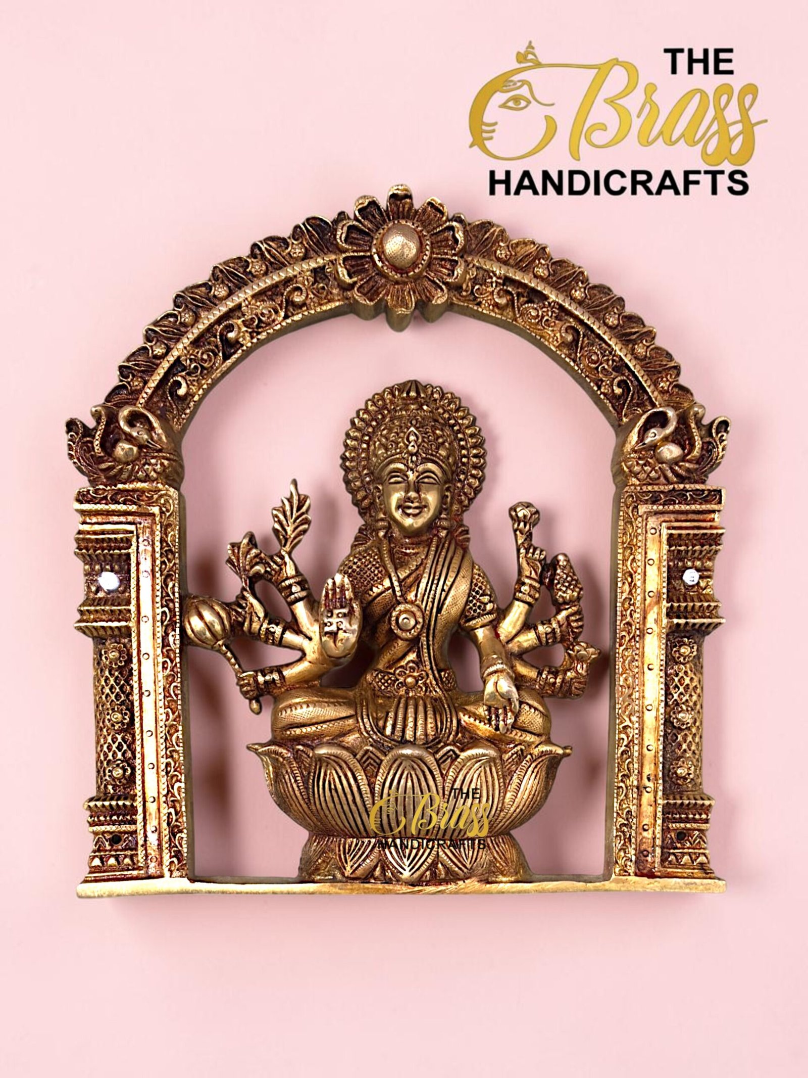 Gold brass statue of a multi-armed Hindu deity seated on a lotus, within an ornate arch against a pink background.  thebrasshandicrafts.com