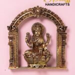 Ornate brass Lakshmi statue seated on a lotus within a decorative arch, pink background, logo 'The Brass Handicrafts' in corner.  thebrasshandicrafts.com