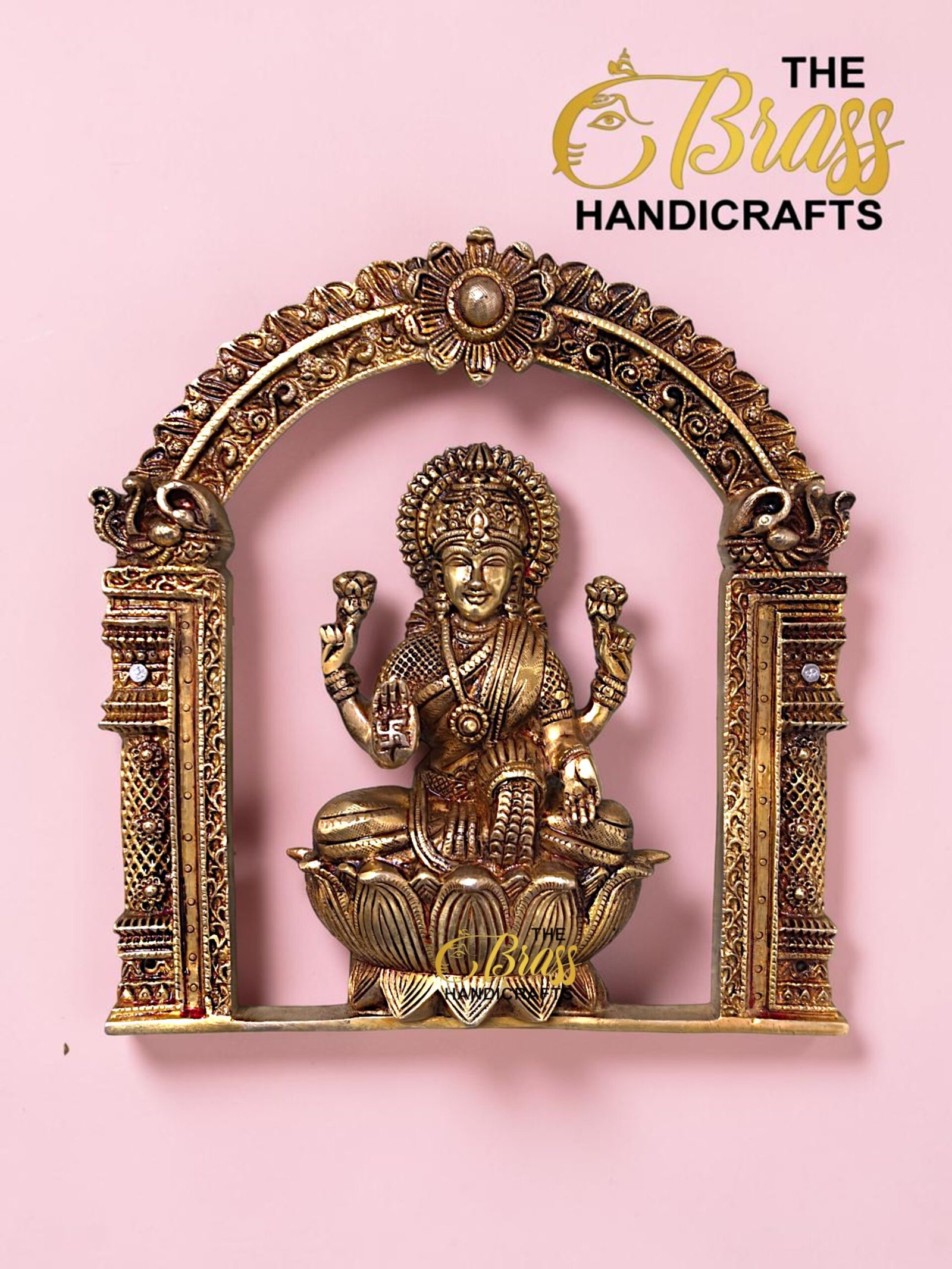 Ornate brass Lakshmi statue seated on a lotus within a decorative arch, pink background, logo 'The Brass Handicrafts' in corner.  thebrasshandicrafts.com