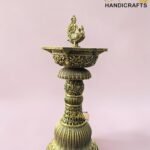 Ornate brass ceremonial vessel with a decorative peacock finial on top, set against a pink background.  thebrasshandicrafts.com