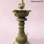 Ornate brass pedestal with a decorative bird finial atop a floral, engraved base against a pink background.  thebrasshandicrafts.com