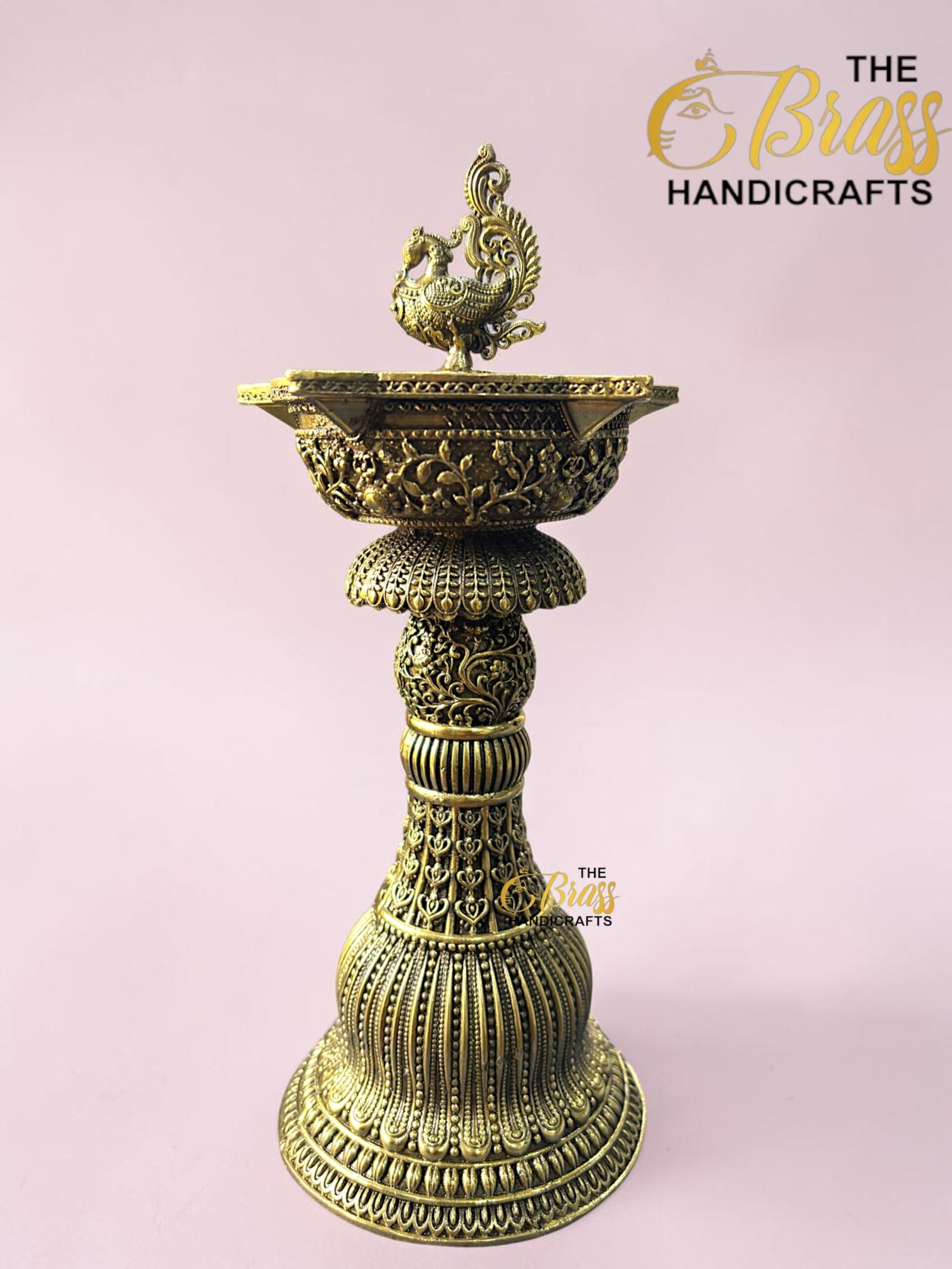 Ornate brass pedestal with a decorative bird finial atop a floral, engraved base against a pink background.  thebrasshandicrafts.com