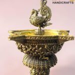 Elaborate brass sculpture featuring a peacock-like bird atop a decorative bowl with intricate filigree patterns against a pink background.  thebrasshandicrafts.com