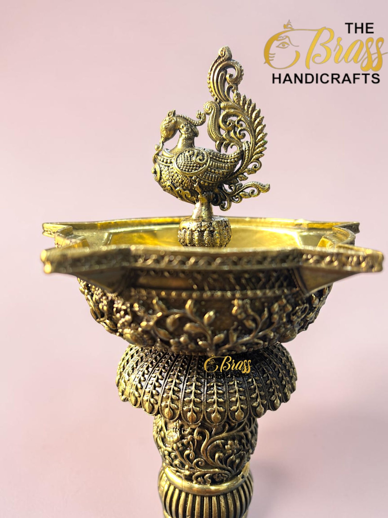 Elaborate brass sculpture featuring a peacock-like bird atop a decorative bowl with intricate filigree patterns against a pink background.  thebrasshandicrafts.com