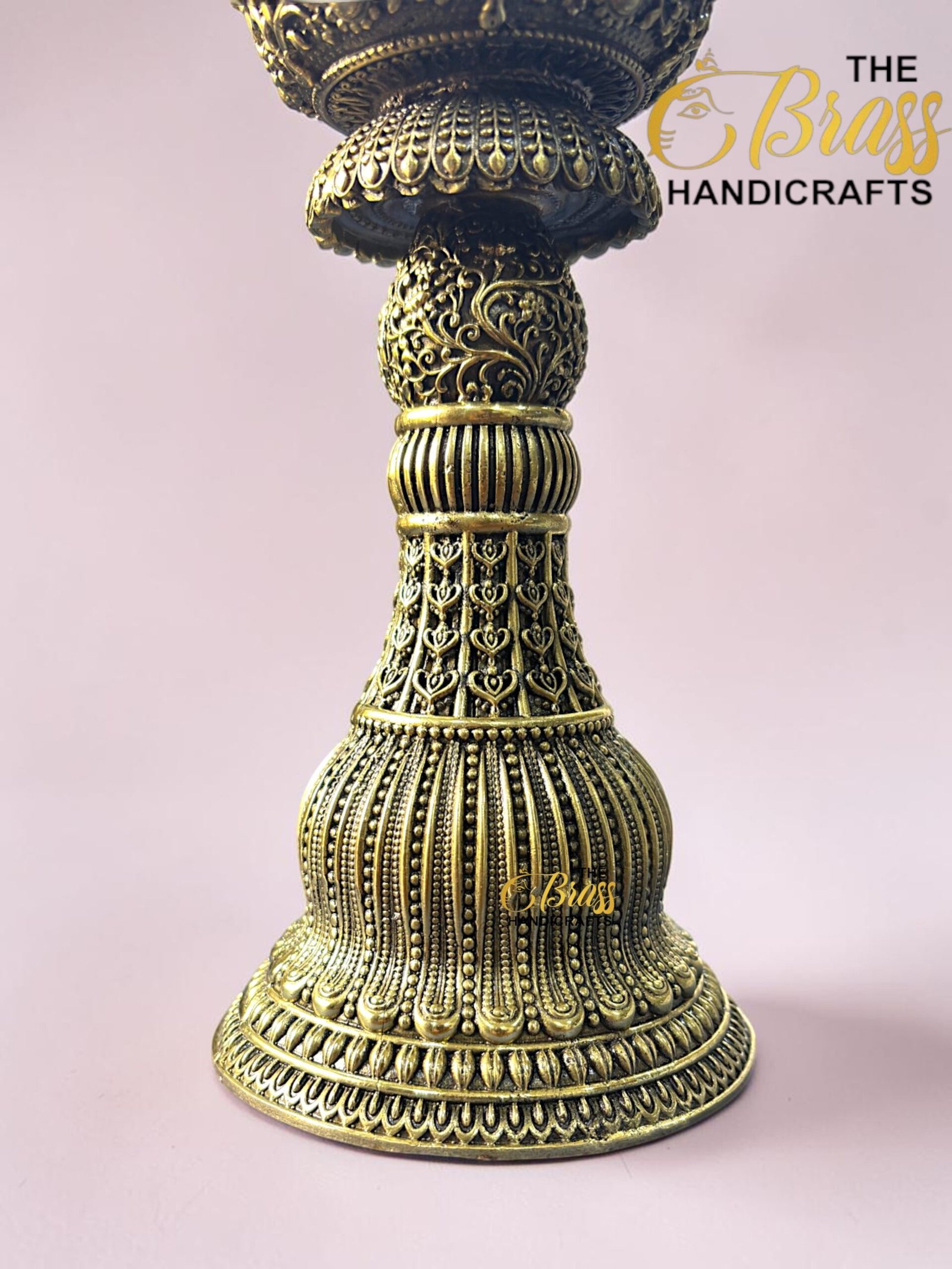 Close-up of an ornate brass pedestal with floral filigree on a pink background; logo reads The Brass Handicrafts in the corner.  thebrasshandicrafts.com