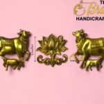 Gold brass cow statues flanking a central lotus ornament on a pink background, with 'The Brass Handicrafts' branding visible.  thebrasshandicrafts.com