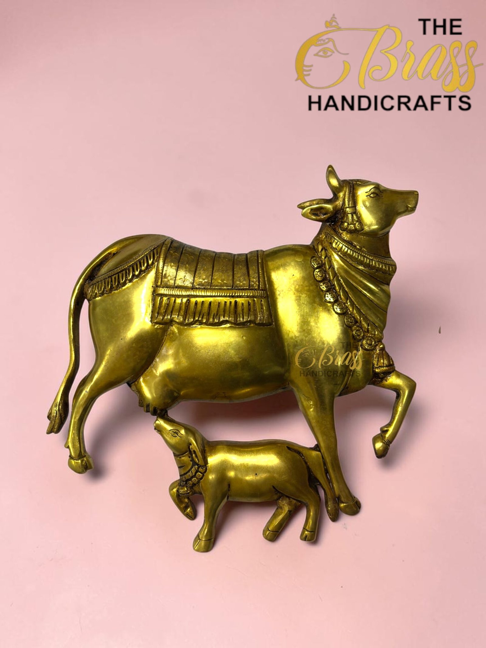 Gold brass statue of a decorated cow with saddle and beaded necklace on a pink background.  thebrasshandicrafts.com