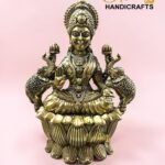 IMG_5773 - thebrasshandicrafts.com Golden brass statue of a Hindu goddess seated on a lotus pedestal with an ornate headdress, set against a pink background with a logo in the corner. thebrasshandicrafts.com